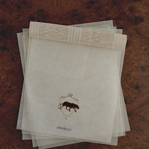 May include: A stack of translucent white paper sheets with a decorative beige trim. Each sheet features a gold emblem of a panther inside a shield, with the text "elmoire.co" below.