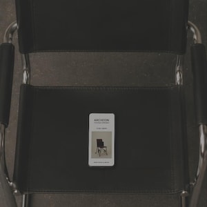 Puede incluir: Una silla de cuero negro con estructura tubular cromada y reposabrazos negros. Sobre el asiento hay una tarjeta rectangular blanca con el texto ARCHEION MOCKUP COLLECTION, 5152 X 3000PX, y una pequeña imagen del mismo diseño de silla.