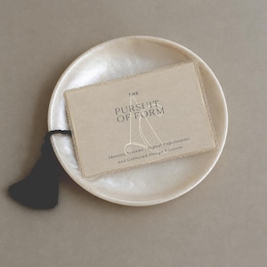 May include: A rectangular beige card with frayed edges and a black tassel, resting on a round, iridescent white dish. The card features the text "THE PURSUIT OF FORM" and "Identity Systems, Digital Experiences, And Collected Design Editions" in black.