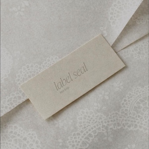 May include: A rectangular label seal with the words "label seal" and "mockup" printed in a light gray color. The label is placed on a light gray paper with a delicate lace pattern.