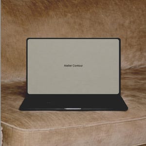 Può includere: Un laptop nero con il testo "Atelier Contour" sullo schermo, appoggiato su un divano di velluto marrone. Il laptop è aperto, mostrando uno schermo beige chiaro. Il divano ha un design trapuntato.