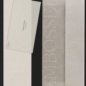 May include: A light-coloured stationery set is shown in a vertical image. The set includes a textured envelope with the word "EMBOSSED" and a matching card with the text "Atelier Contour" and "Your Design Here". The stationery is a neutral colour.