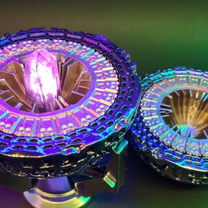 Könnte beinhalten: Zwei futuristische, irisierende Objekte mit aufwendigen Designs. Eines hat einen zentralen, leuchtenden lila Kristall. Das andere ist ein ähnliches, kleineres Design. Die Objekte haben eine metallische, mehrfarbige Oberfläche, die ein Sci-Fi- oder Fantasy-Thema suggeriert.