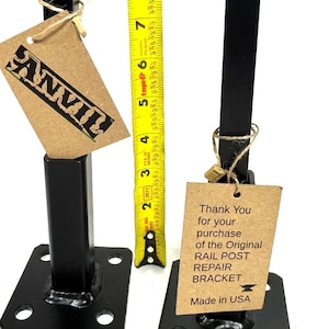 May include: Two black metal rail post repair brackets with a square base and four holes. Each bracket is attached to a brown tag with text. The tag on the left bracket says "ANVIL". The tag on the right bracket says "Thank You for your purchase of the Original RAIL POST REPAIR BRACKET Made in USA". A yellow tape measure is visible in the background.