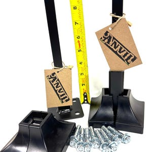 May include: Two black metal fence post bases with square tops and a decorative design. Each base is attached to a black metal post with a brown paper tag that says "ANVIL". The bases are sitting on a white surface with a set of silver lag screws.