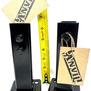 May include: Two black metal brackets with bolt fasteners and a square base. Each bracket has a tag with the word "ANVIL" printed on it. A yellow tape measure shows the height of the brackets is approximately 8 inches.