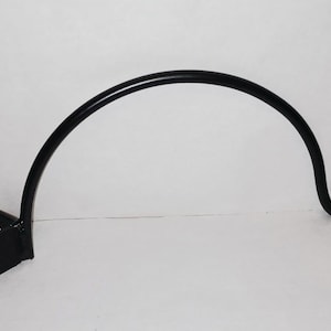 May include: Black metal plant hanger with a curved, arched design. The hanger features a bracket with two holes for mounting and a curved hook at the end. The overall design is simple and functional.