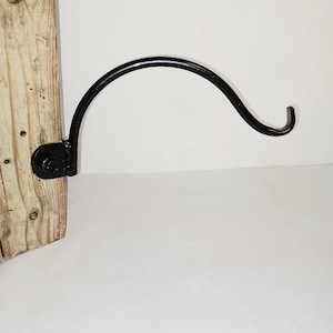 May include: A black, curved metal hook attached to a piece of weathered, light-colored wood. The hook is designed to hold items and is mounted with a black metal plate and screws. The wood has visible knots and a rustic appearance.