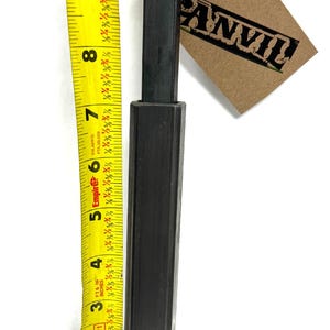 May include: A black metal rod with a square cross-section, measuring approximately 7 inches in length. The rod is tied with twine and has a brown paper tag with the word "ANVIL" printed on it. A yellow measuring tape is visible in the background.