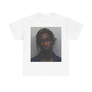 Puede incluir: Camiseta blanca con una imagen rectangular de una persona con cabello castaño claro y una expresión seria. La persona lleva una camisa azul oscuro. El fondo es gris claro.