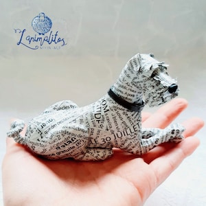 Puede incluir: Una escultura de papel maché blanca de un perro acostado. La escultura está hecha de papel reciclado y está cubierta de texto de un periódico. El perro lleva un collar negro.