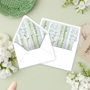 May include: Two white envelopes with floral patterned inserts. The inserts feature vertical stripes in shades of green and blue with botanical accents. The envelopes are on a light green surface, with white flowers and decorative elements in the background.