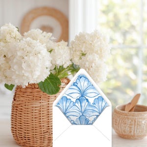 May include: An open white envelope with a blue floral pattern inside, displayed in front of a wicker basket filled with white flowers. A small, decorative ceramic bowl is in the background.