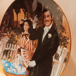 May include: A decorative ceramic plate featuring a couple in a vintage setting. The man is wearing a black suit and top hat, while the woman is wearing a striped dress and a hat. The plate has a gold rim and a black background.
