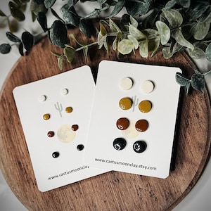 May include: Two sets of clay stud earrings on white cards. The first set includes two white, two mustard yellow, two brown, and two black earrings. The second set includes two white, two mustard yellow, and two brown earrings. The cards have the website address "www.cactusmoonclay.etsy.com" printed on them.
