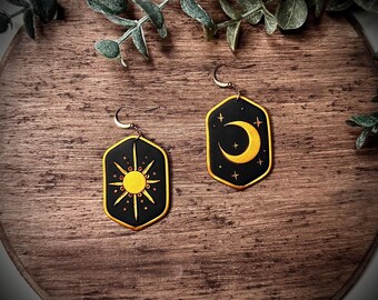 May include: A pair of black and gold earrings with a sun and moon design. The earrings are shaped like a hexagon and have a gold outline. The sun is in the center of one earring and the moon is in the center of the other earring.