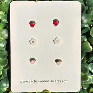 May include: A set of six polymer clay earrings featuring three pairs of miniature strawberries and white flowers. The earrings are displayed on a white card with the text "www.cactusmoonclay.etsy.com".