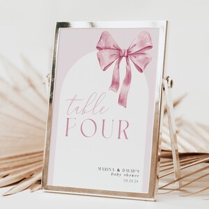 Könnte beinhalten: Ein rechteckiger goldfarbener Metallrahmen mit einem Tischschild, das einen hellrosa Hintergrund, eine weiße Bogengrafik und eine rosa Aquarellschleifen-Illustration zeigt. Der Text lautet "table FOUR" und "MARINA & DAVID'S baby shower 26.08.24".