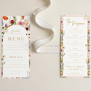 May include: Two floral-themed wedding stationery cards. One is a menu with an arched top, the other a program. Both have a white background with gold text and colorful flower borders. A white ribbon is draped across the top.