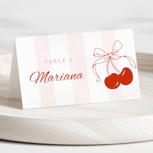 May include: A white table card with pink and white stripes. The text "TABLE 1 Mariana" is printed in red. A red cherry with a bow is illustrated on the right side of the card.