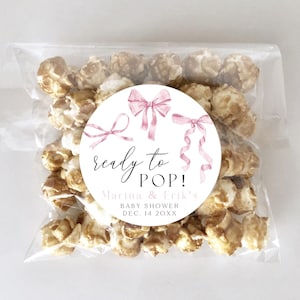 Puede incluir: Una pegatina circular blanca con lazos de acuarela rosa y el texto "ready to POP!"  "Marina & Erik's Baby Shower Dec. 14 20XX"  La pegatina está en una bolsa de plástico transparente llena de palomitas de maíz.