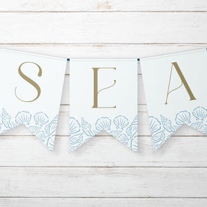 May include: A banner with white pennants spelling "SEA" in gold letters. Each pennant has a blue seashell design along the bottom edge. The banner is strung on a blue string and set against a white wooden background.