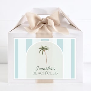 Puede incluir: Caja de regalo blanca con una cinta beige y una etiqueta a rayas azules y blancas. La etiqueta tiene una ilustración de una palmera y el texto "Jennifer's Beach Club".