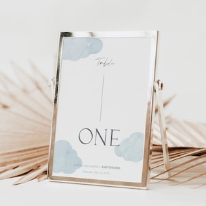 May include: A silver picture frame with a white card inside. The card has a blue watercolor cloud design and the text "Table One" and "Alison and Gabriel Baby Shower Saturday, July 23, 2023".