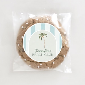 May include: A clear plastic bag with white polka dots contains a chocolate chip cookie with a round sticker. The sticker has a blue and white striped background with a palm tree and the text "Jennifer's Beach Club".