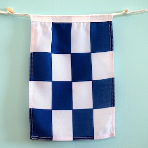 Könnte beinhalten: Ein rechteckiges Stoffbanner mit einem sich wiederholenden Schachbrettmuster aus abwechselnden königsblauen und weißen Quadraten. Das Banner ist an einer weißen Kordel mit kleinen Holzperlen an den Enden befestigt und hat einen genähten Saum.
