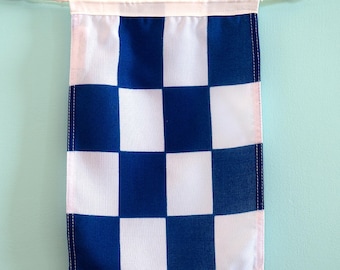 May include: A rectangular fabric banner featuring a repeating chequered pattern of alternating royal blue and white squares. The banner is suspended from a white cord with small wooden beads at each end, featuring a clean stitched top hem.