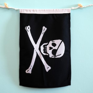 May include: A rectangular black fabric wall banner featuring a white screen-printed graphic of a skull with an eye patch and two crossed bones. The banner has a white fabric header at the top with a white cord threaded through for hanging.