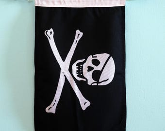May include: A rectangular black fabric wall banner featuring a white screen-printed graphic of a skull with an eye patch and two crossed bones. The banner has a white fabric header at the top with a white cord threaded through for hanging.