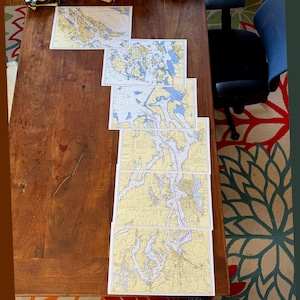 May include: A collection of nautical charts, featuring detailed illustrations of coastal areas with blue, yellow, and white color schemes. The charts are arranged on a dark wooden table, showcasing various geographical features.