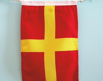 May include: A rectangular fabric wall banner featuring a bold yellow cross centered on a vibrant red background. The banner is suspended from a white cord with small wooden toggles at each end, attached to a white fabric header at the top edge.