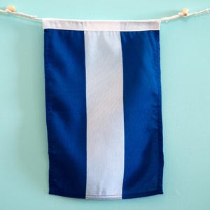Op de afbeelding: Een rechthoekige stoffen wimpel met een verticaal driekleurig ontwerp met twee donkerblauwe buitenpanelen en een centraal wit paneel. De vlag hangt aan een witte horizontale zoom die is bevestigd aan een dun wit koord met kleine houten kralen.