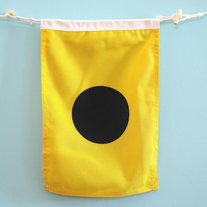 May include: A rectangular yellow nautical signal flag featuring a large solid black circle in the center. The flag has a white fabric header at the top with white rope and wooden toggles on each side for hanging, displaying a clear maritime design.