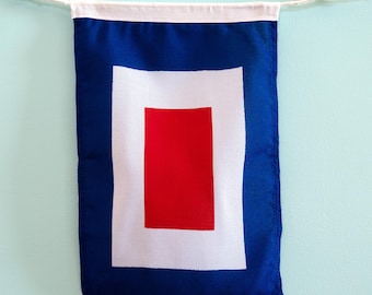 May include: A rectangular nautical signal flag featuring a solid blue border, a white rectangular frame, and a solid red rectangular center. The flag is suspended from a white rope with wooden toggles at the top, displaying a classic maritime design.