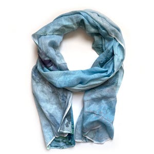 May include: A light blue scarf with a watercolour-like pattern of blue, green, and white. The scarf is made of a soft, lightweight fabric and has a fringed edge.