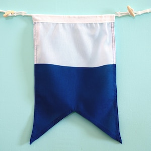 May include: A rectangular fabric pennant flag featuring a horizontal color-blocked design with a white upper section and a deep blue lower section. The bottom edge is cut into a pointed V-shape. It hangs from a white cord with wooden toggles.