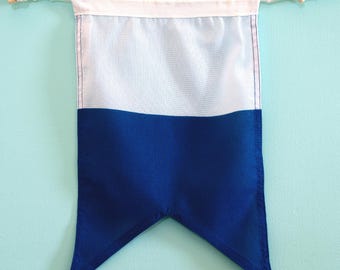 May include: A rectangular fabric pennant flag featuring a horizontal colour-blocked design with a white upper section and a deep blue lower section. The bottom edge is cut into a pointed V-shape. It hangs from a white cord with wooden toggles.