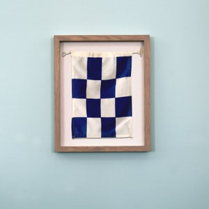 May include: A framed nautical signal flag with a white background and blue checkered pattern.