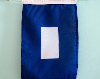 May include: A rectangular blue fabric wall banner featuring a centred vertical white rectangle. The banner is suspended from a white cord with small wooden toggles at each end, creating a clean and simple display piece for wall decoration.