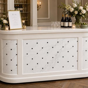 Könnte beinhalten: Eine weiße Bar mit marmorähnlicher Front, verziert mit schwarzen quadratischen Akzenten. Die Bar verfügt über ein goldgerahmtes Schild, Weinflaschen, Gläser und Blumengestecke. Die Bar ist für eine besondere Veranstaltung eingerichtet.