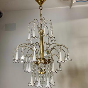 Op de afbeelding: Een vintage kroonluchter met een messing afwerking en trapsgewijze kristallen accenten. De armatuur heeft witte, klokvormige kappen met zwarte rand. Het ontwerp omvat meerdere gebogen armen en een centrale kolom.