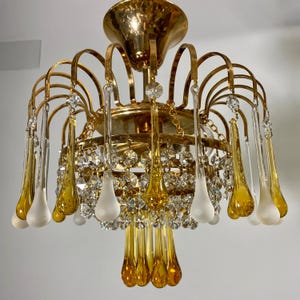 May include: A gold chandelier with clear and amber glass teardrop crystals. The chandelier has a circular base and multiple arms that extend outward, each ending in a teardrop crystal.