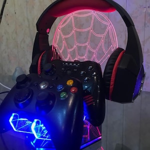 May include: Black gaming controllers with colorful buttons and a black headset with red accents are displayed. A neon pink spiderweb design is in the background, and the base has blue and pink lights.