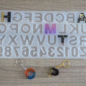 Könnte beinhalten: Eine transparente Kunststoffform mit Alphabet und Zahlen, sowie vier Schlüsselanhänger mit Buchstaben und Zahlen aus Harz. Die Buchstaben sind schwarz mit goldenen Glitzer, und die Zahlen sind in verschiedenen Farben.
