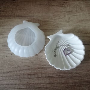 Könnte beinhalten: Zwei weiße Muschelschalen-Schmuckschalen. Eine Schale ist offen, die andere geschlossen. Eine silberne Halskette mit einem herzförmigen lila Edelstein liegt in der offenen Schale. Die Schalen stehen auf einer Holzoberfläche.
