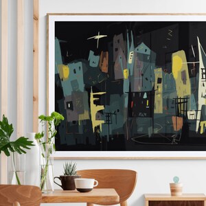 May include: Abstract cityscape art print in a wooden frame. The artwork features a dark background with stylized buildings in shades of teal, yellow, and brown. A star-like shape is visible in the upper left corner. The print is displayed on a wall above a wooden table.
